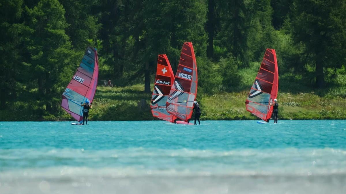 Group of windsurfers on a turquoise lake bordered by lush forest and hills.