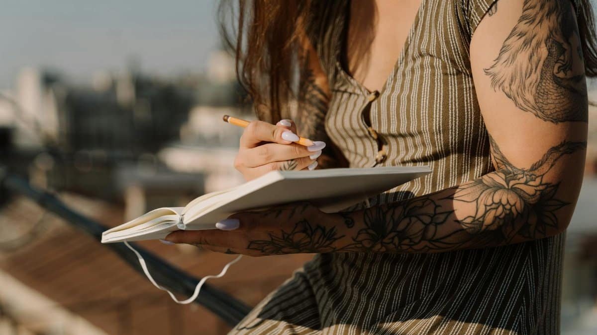 A young woman with tattoos writes in a notebook while sitting on a sunny urban rooftop.