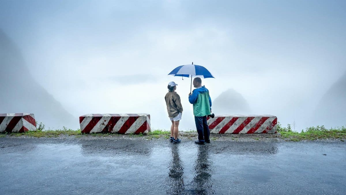 A couple stands on a wet road under an umbrella near concrete barriers with a misty view.