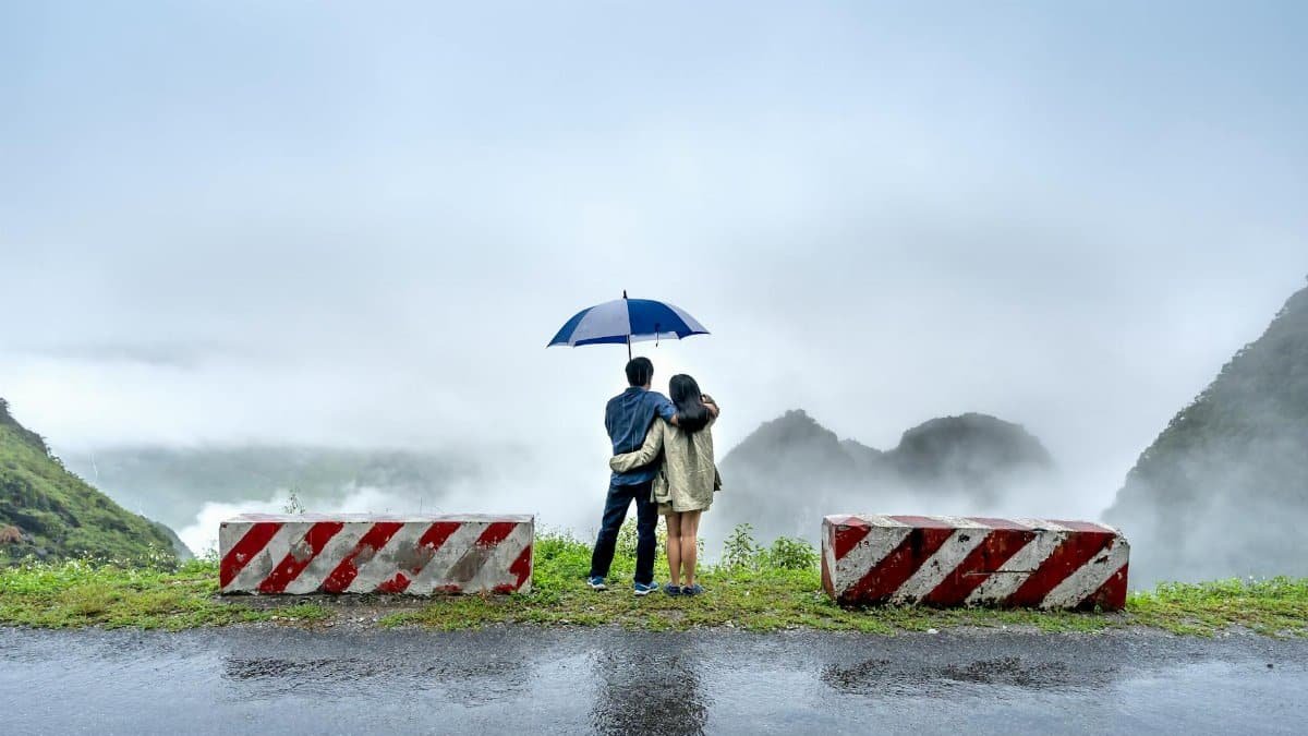 A couple stands under an umbrella, gazing at a foggy landscape from a cliff with wet roads and barriers.