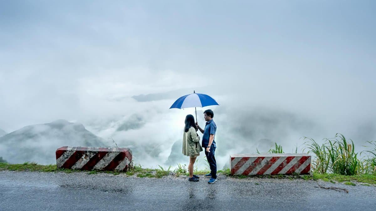 A couple standing with an umbrella on a misty cliff road surrounded by concrete barriers.