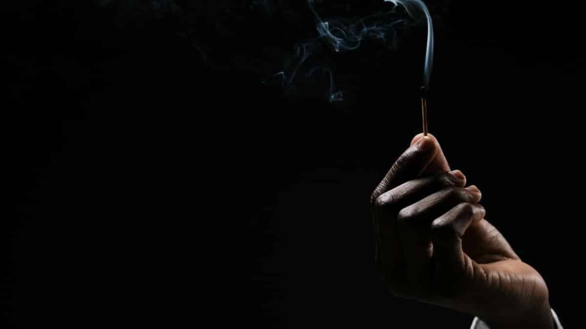 A hand holds a lightly smoking match against a dark background, capturing a dramatic and moody feel.