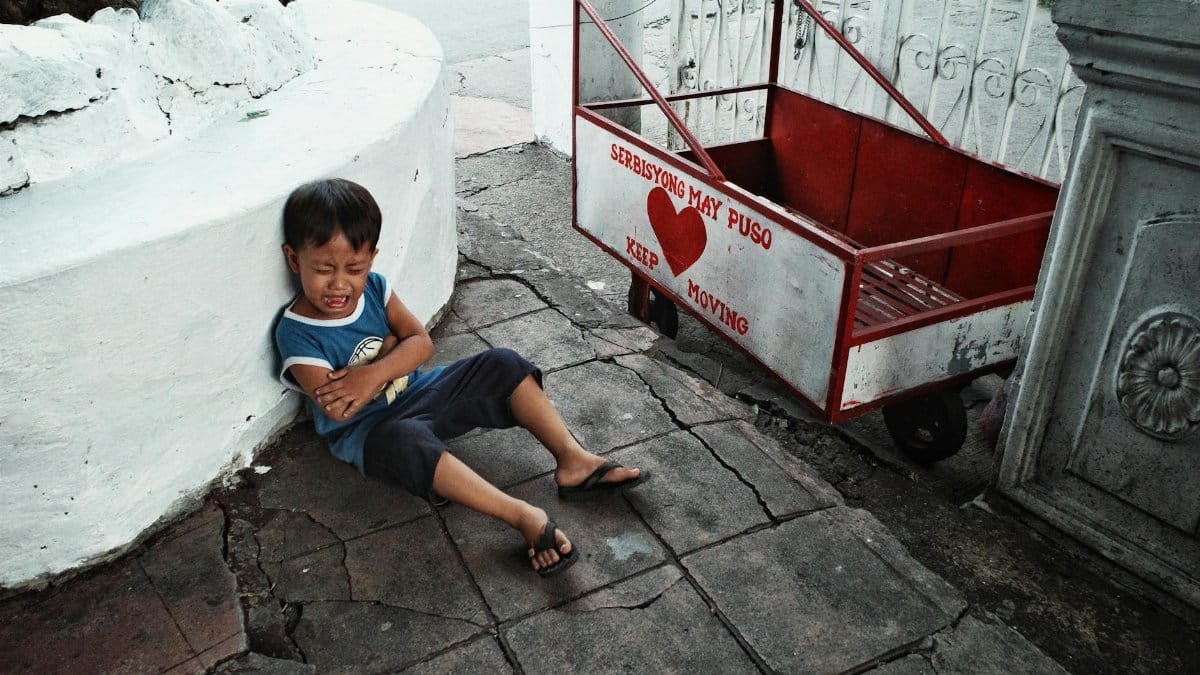 Candid street photograph of a crying child sitting on a city sidewalk near a red cart.