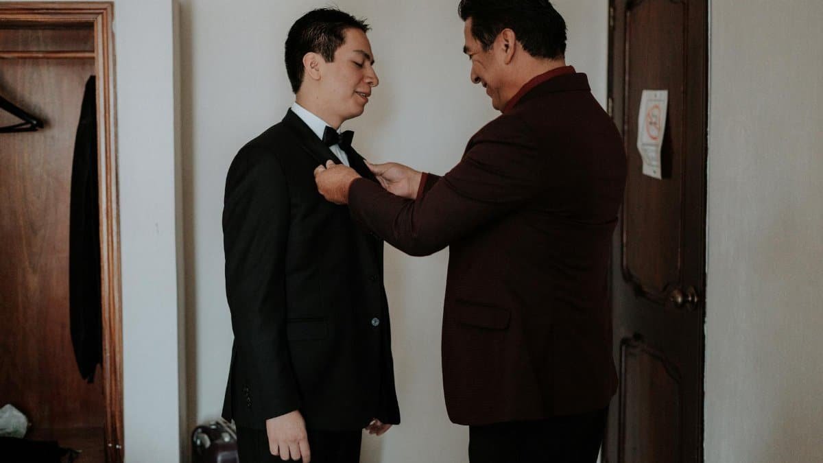 Father assists son with bow tie, creating a heartwarming moment in formal attire setting.