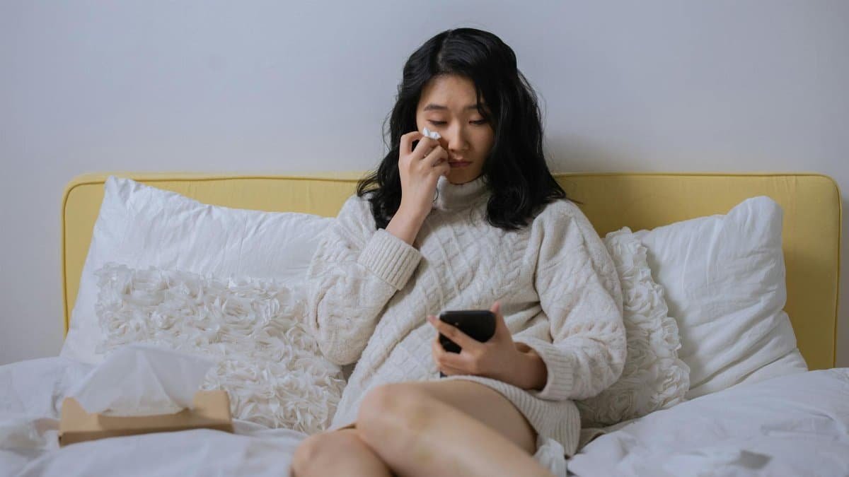 Asian woman in a sweater, looking sad and wiping tears, holding a smartphone seated on a bed.