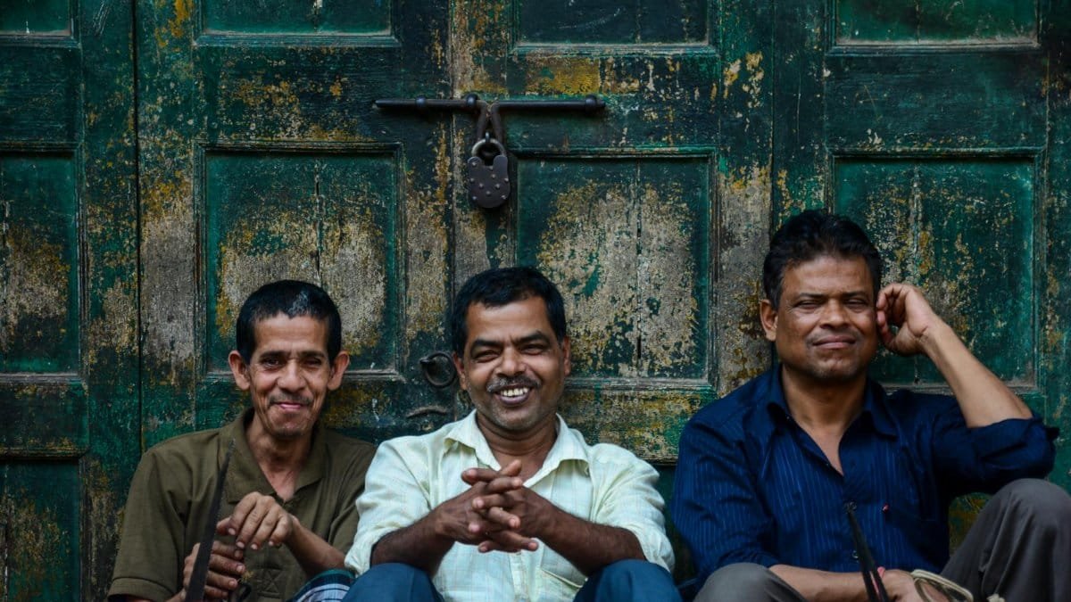 Three men sitting and smiling against a weathered wooden door outdoors.
