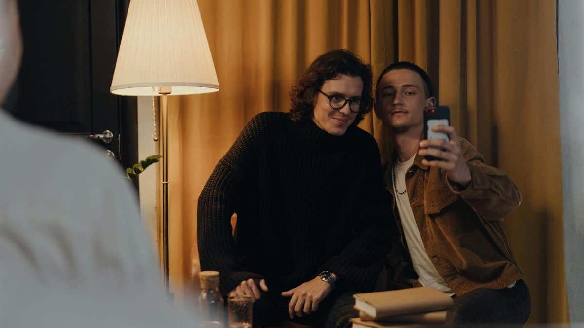 Two young men capture a selfie indoors with books stacked nearby, creating a cozy ambiance.