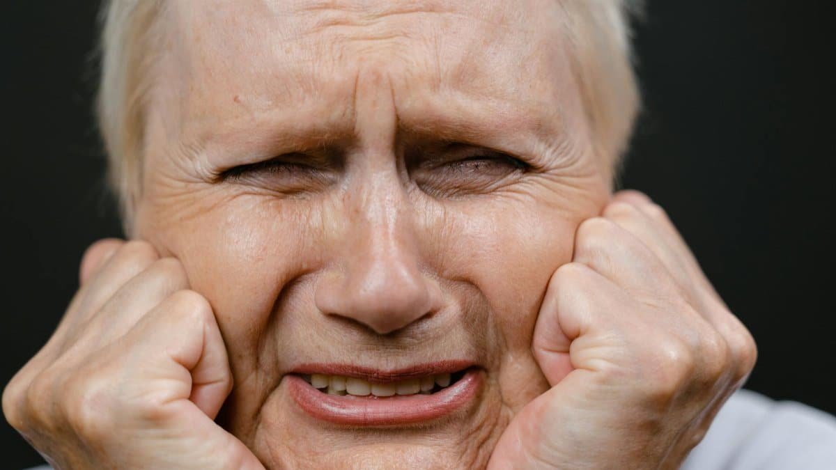 Close-up of a distressed elderly woman expressing deep emotion with hands on face.