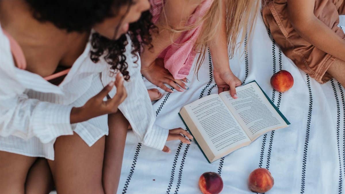 Teenage friends enjoying a sunny picnic with a book and peaches on a striped blanket.