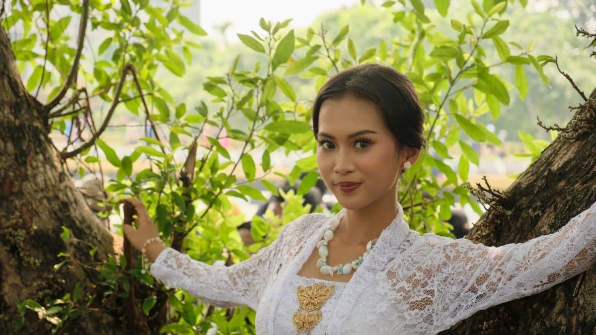 A woman in traditional Indonesian attire poses elegantly amid lush greenery outdoors, capturing cultural beauty.