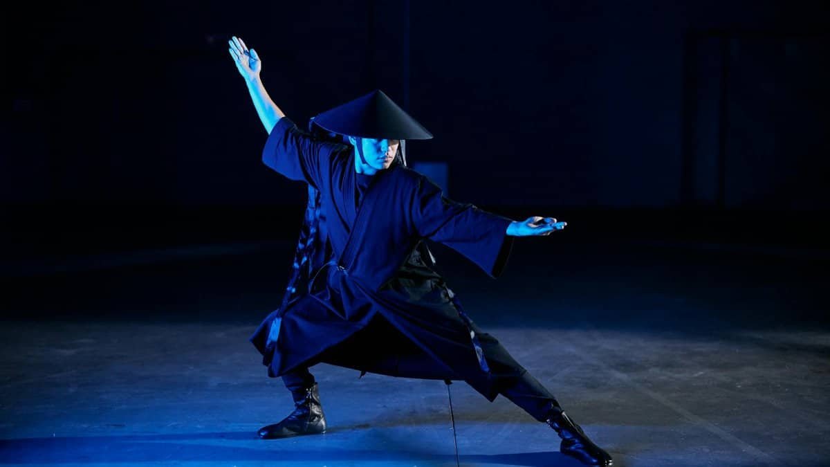 A martial artist dressed in traditional attire strikes a dynamic pose in an atmospheric indoor setting.