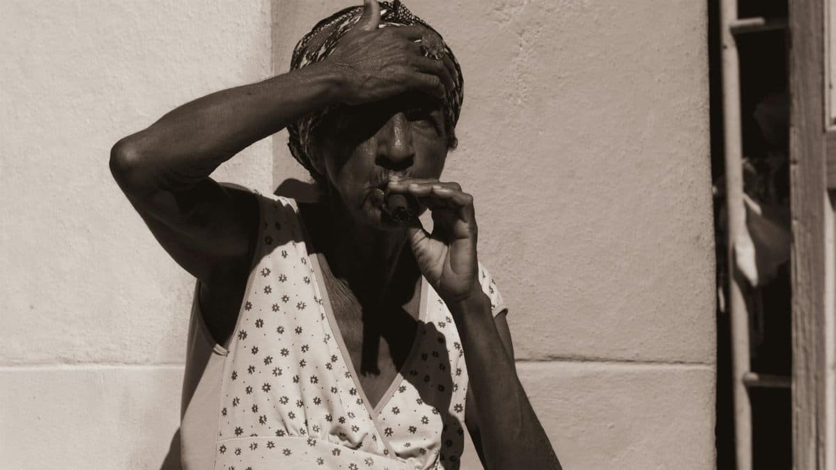 Elderly woman leans against wall, smoking cigar in grayscale, highlighting lifestyle habits.