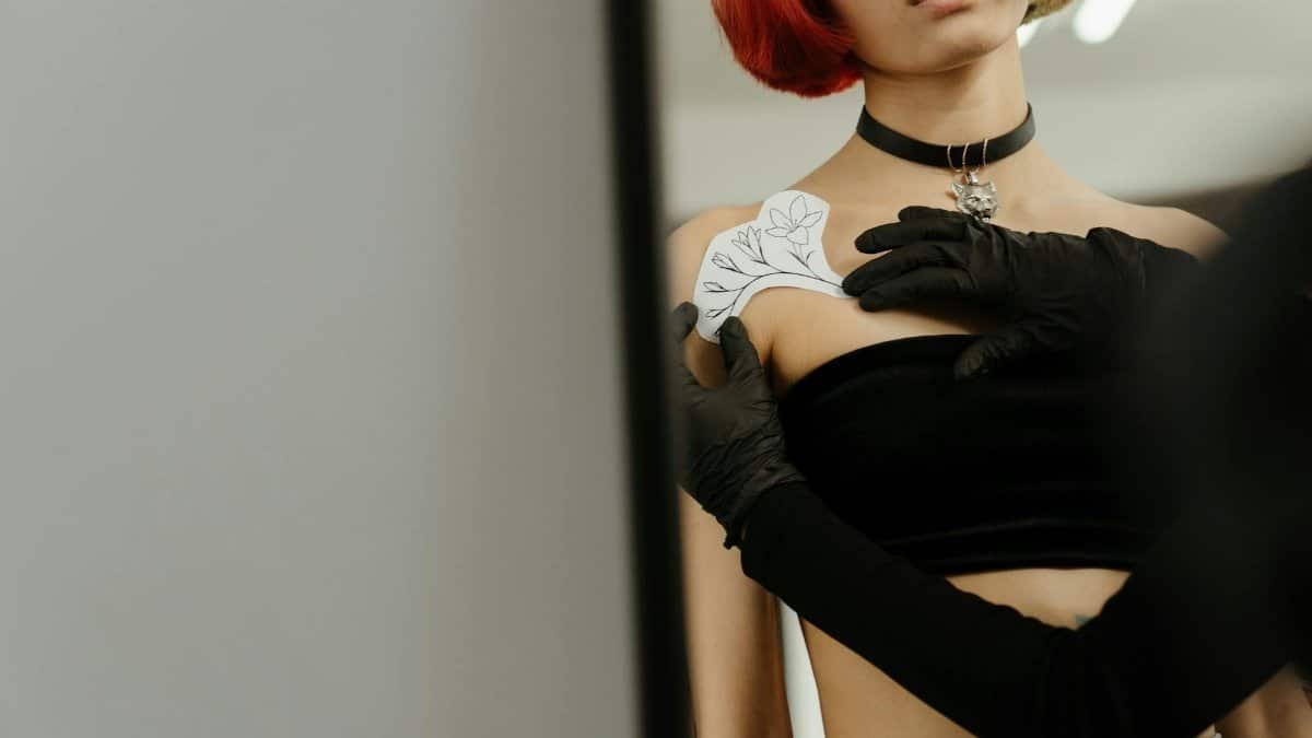 A woman with colored hair getting a temporary tattoo applied on her shoulder at a tattoo studio.