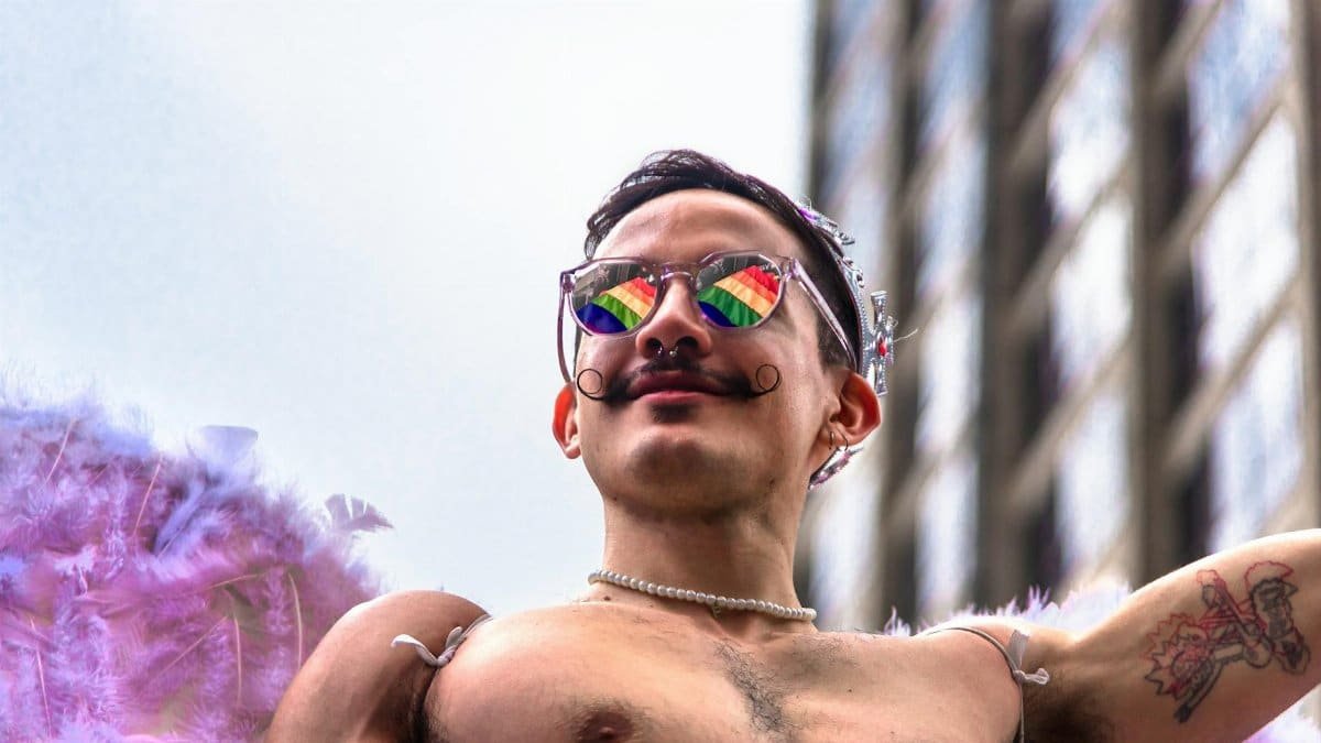 Man with rainbow sunglasses and feathers celebrates at an outdoor pride parade.