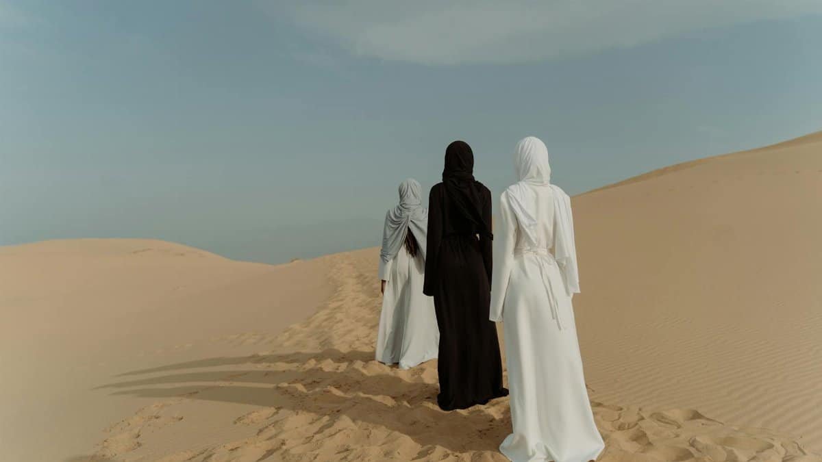 Three women in hijabs and long dresses walking through sand dunes under clear sky.