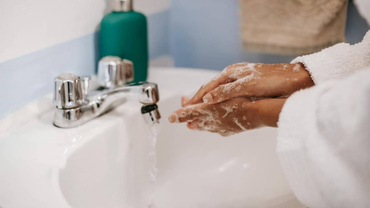 A person in a bathrobe washing hands with soap in a bathroom sink, emphasizing hygiene.