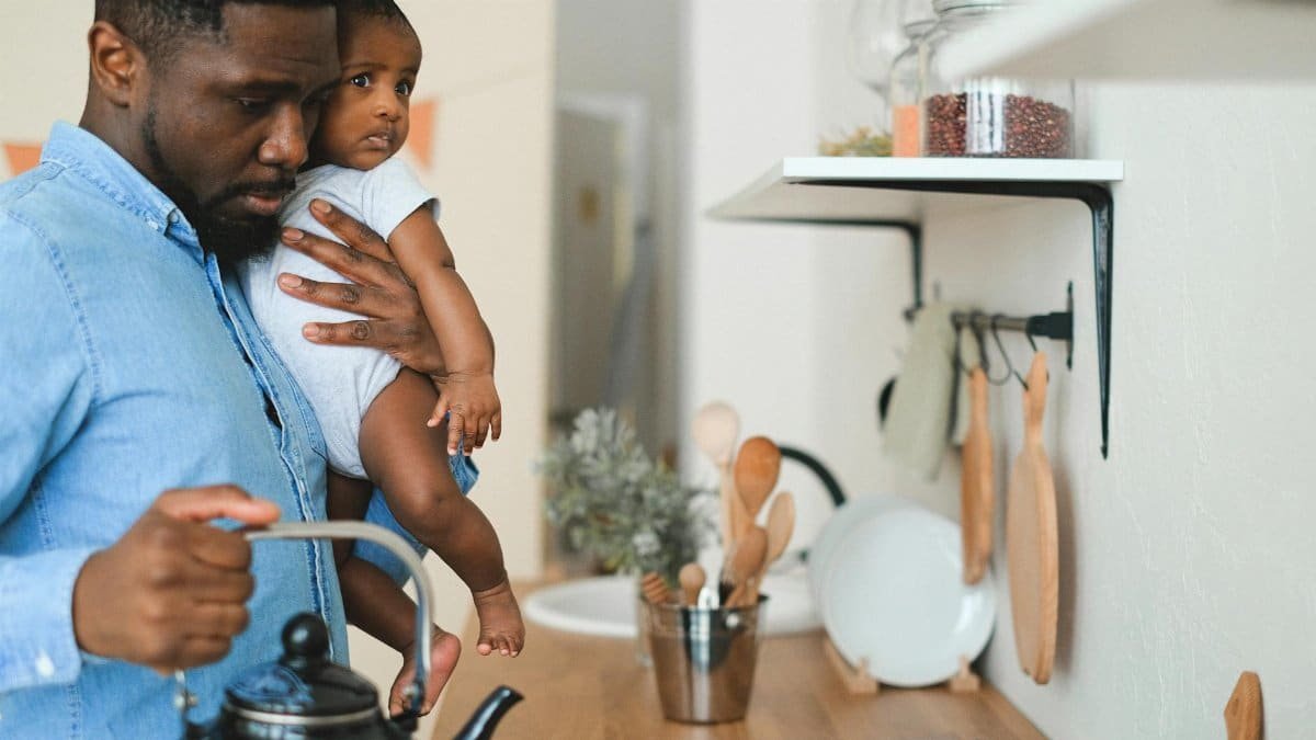 Father holding baby while pouring water from kettle in a modern kitchen setting.