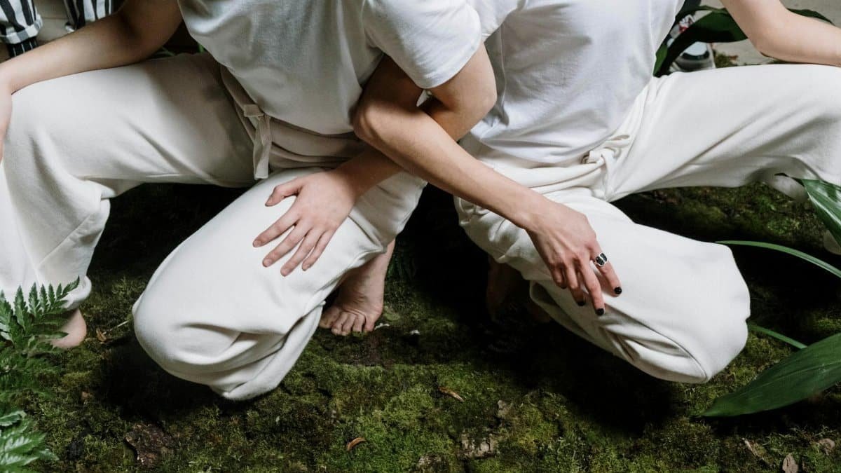 Two people wearing white outfits crouch on a lush, moss-covered ground, outdoors, in a serene setting.