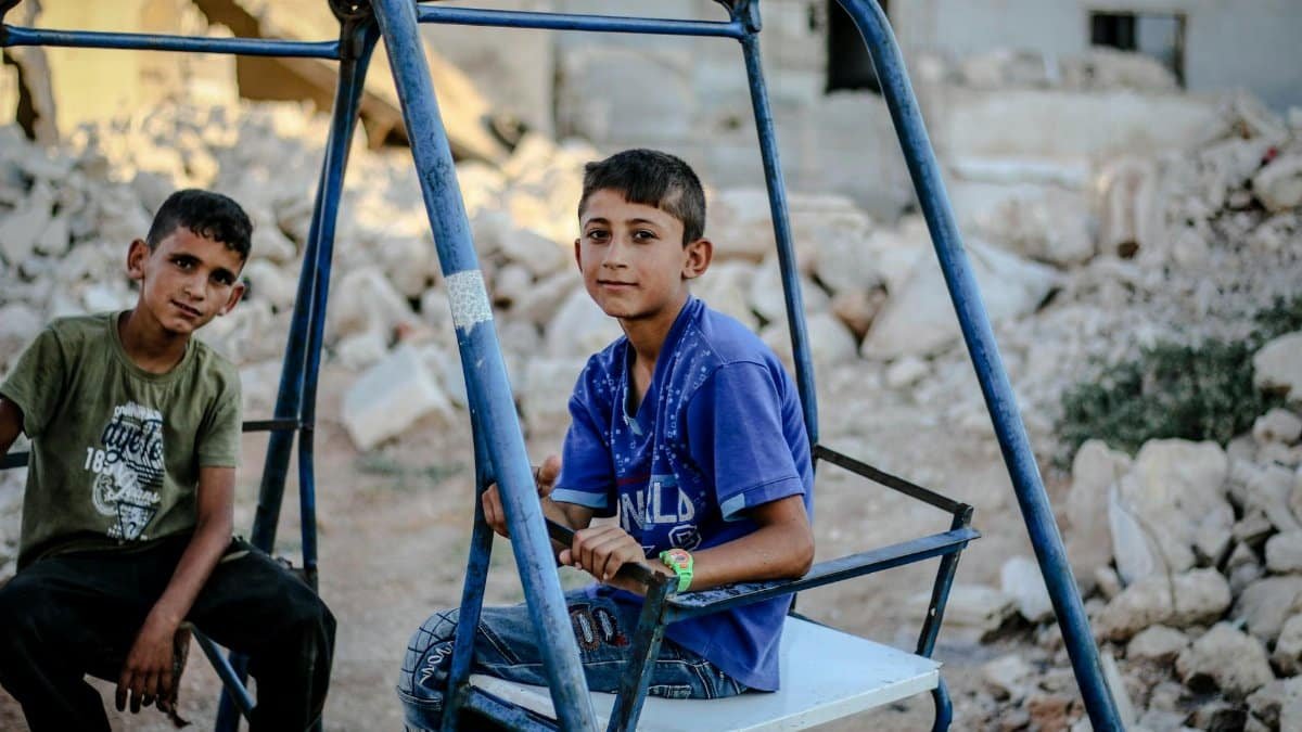 Two boys sitting on a swing outdoors amidst rubble in Idlib, Syria, depicting life resilience.