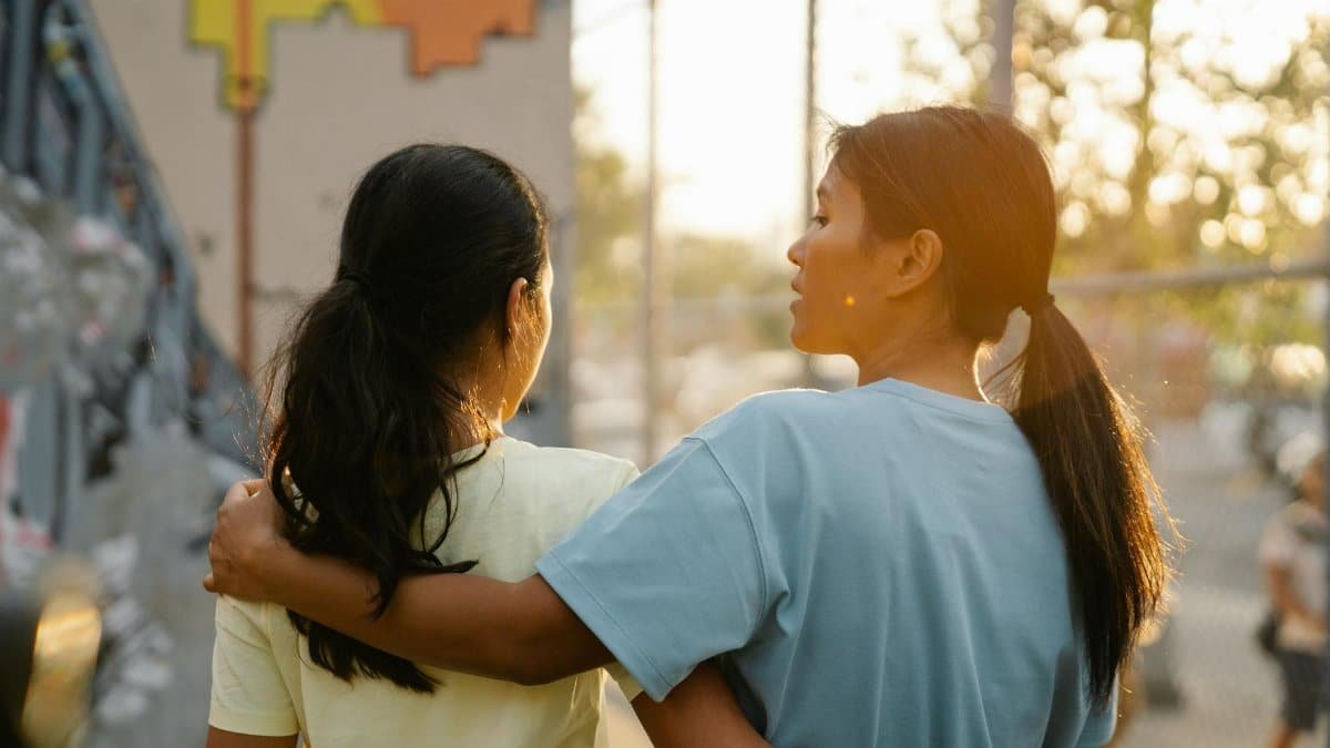 Two women with black hair in ponytails embracing and walking outdoors at sunset, fostering connection.