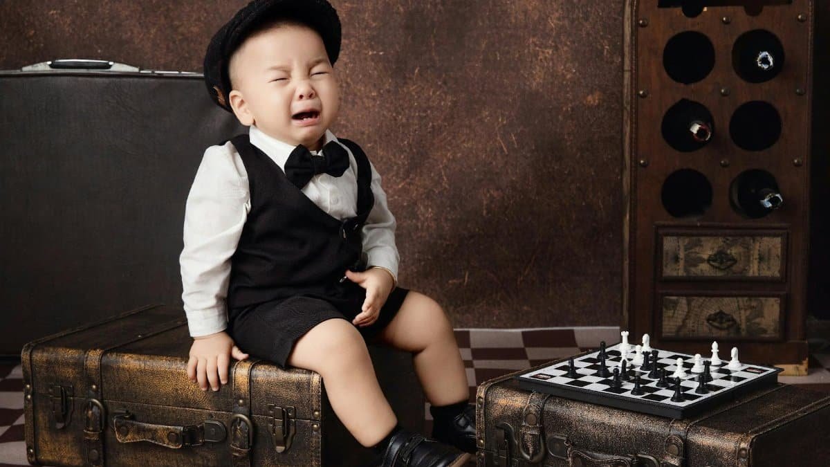 A young boy in formal attire cries while seated on vintage luggage, near a chess board.