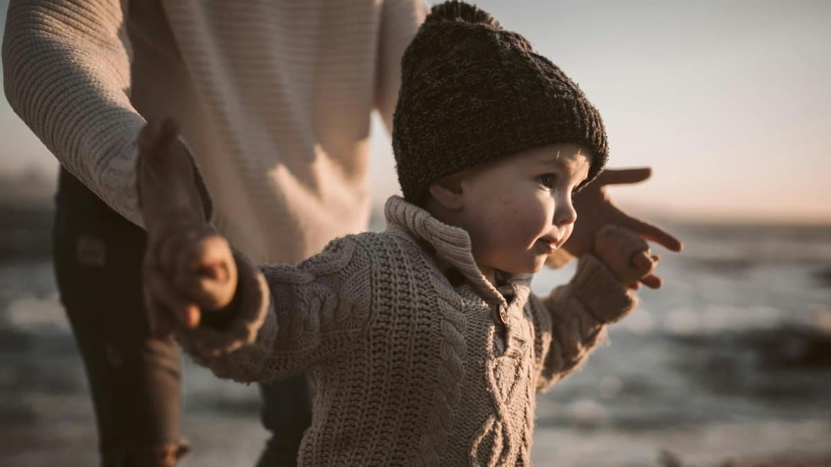 Adorable toddler in a knitted sweater and beanie enjoying a beach walk at sunset, holding hands with an adult.