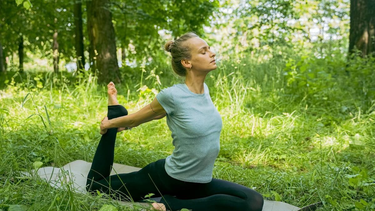 A young woman performs a yoga pose on a mat in a lush green outdoor setting, embracing flexibility and mindfulness.