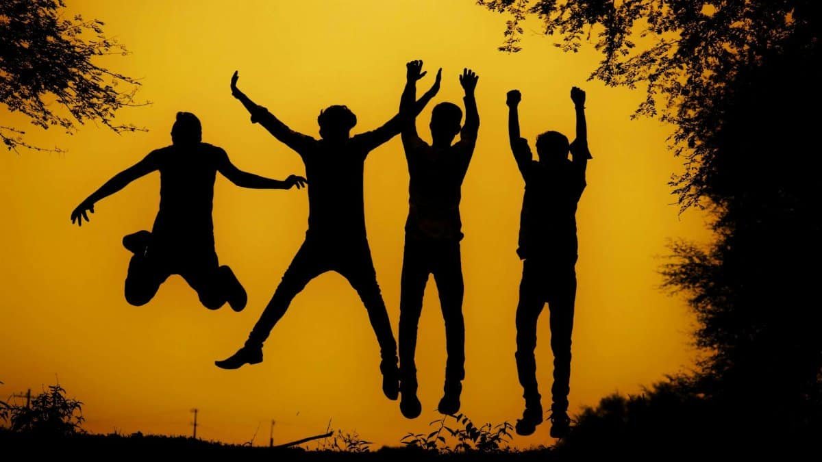 Group silhouetted jump against a vibrant yellow sunset backdrop among trees.