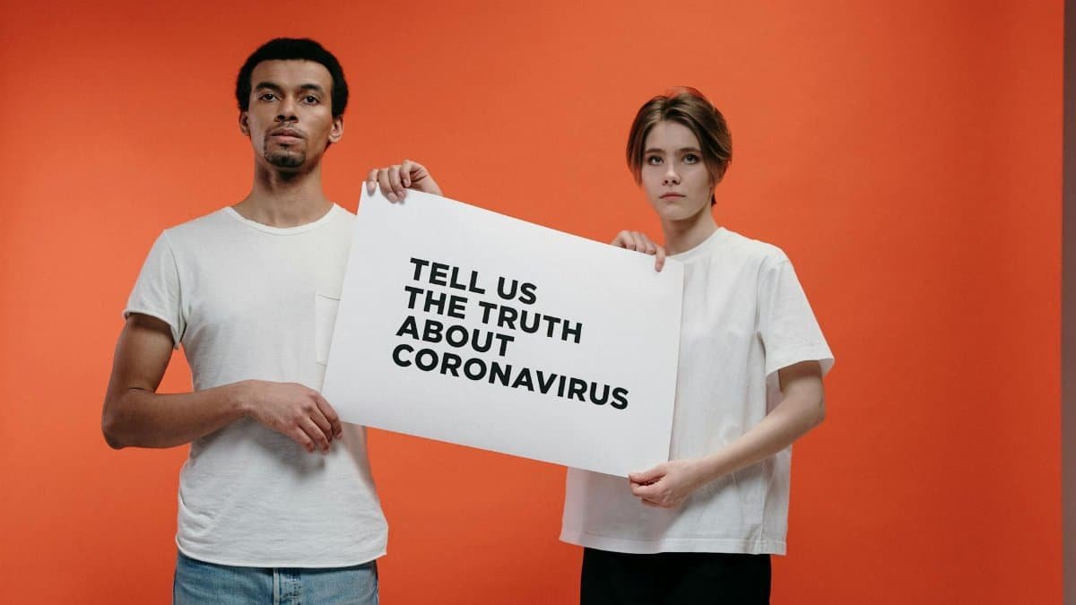 Two people holding a 'Tell Us The Truth About Coronavirus' sign against a vibrant orange background.