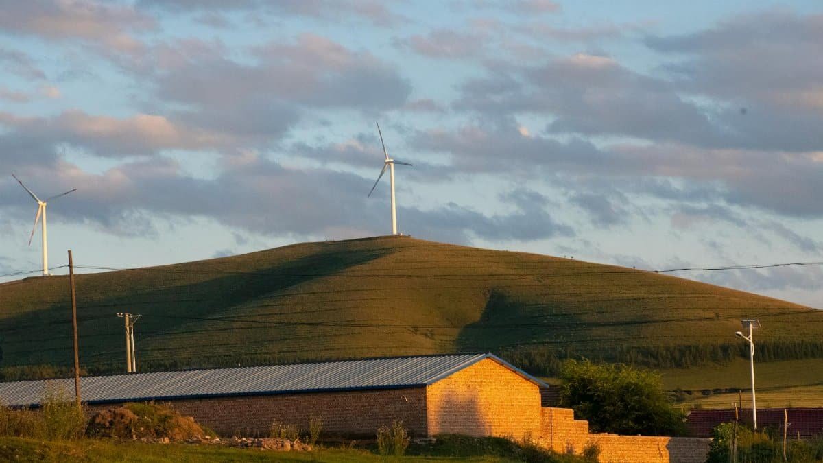 Wind turbines and house on grassy hill during sunset, showcasing renewable energy and rural landscape.