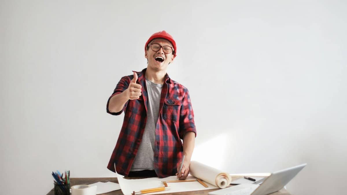 Cheerful male architect in a red hardhat and plaid shirt giving thumbs up at his desk.