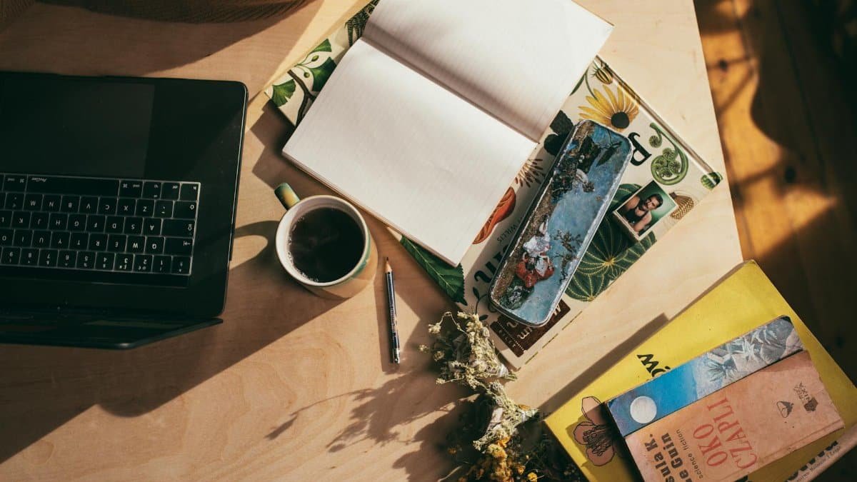 A cozy workspace with an open notebook, laptop, books, and a coffee cup on a wooden table.
