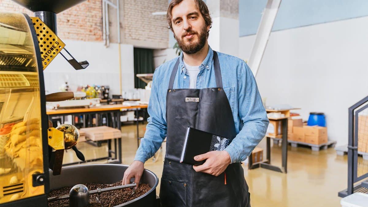 A bearded master roaster stands by a coffee roasting machine, holding a notebook in an industrial workshop.