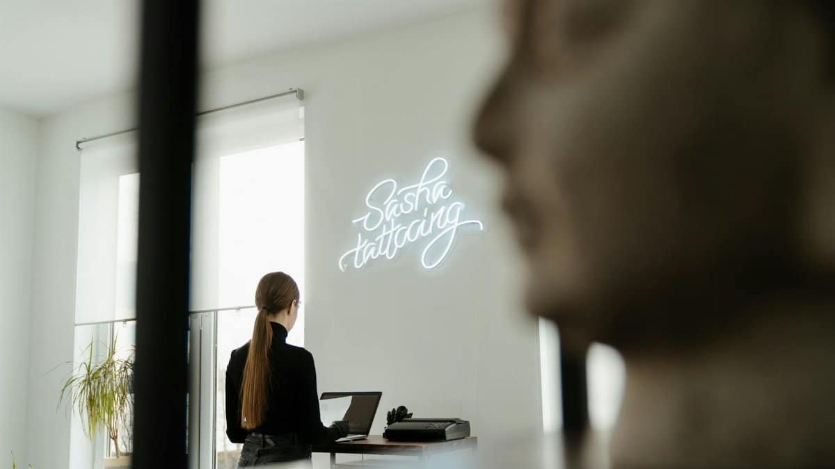 A tattoo artist in a stylish studio with neon sign and natural light.