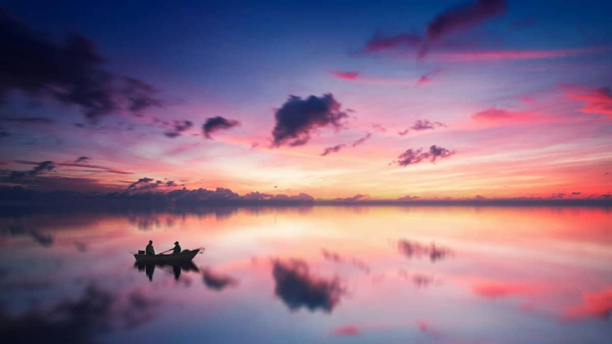 A tranquil sunset scene with a boat silhouetted against a vibrant sky reflected on a calm lake.