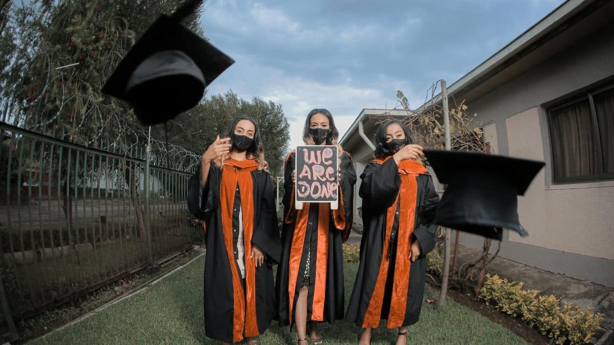 Three graduates in gowns celebrate by tossing caps outdoors, holding 'We Are Done' sign.