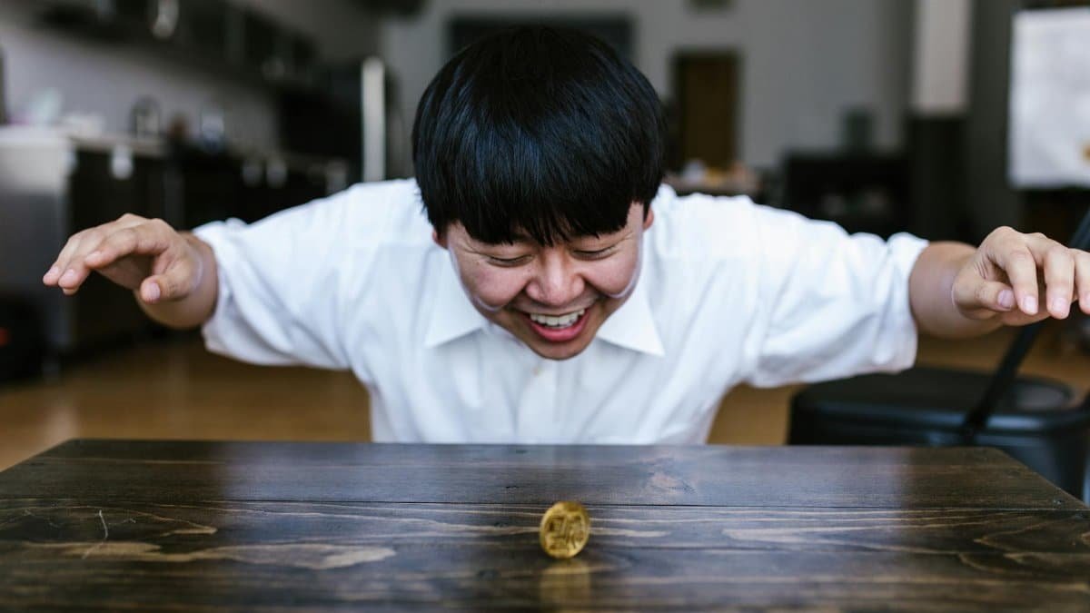 A happy man smiles as he watches a spinning bitcoin coin indoors on a wooden table.