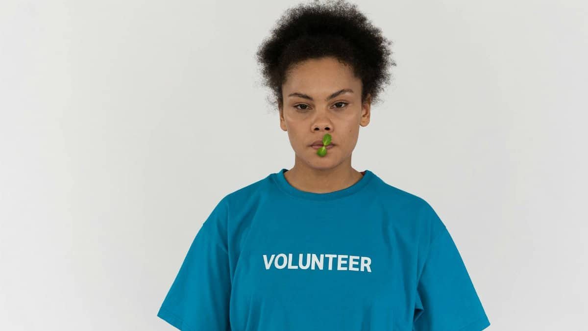 A young woman wearing a volunteer t-shirt holds a plant stem in her mouth.