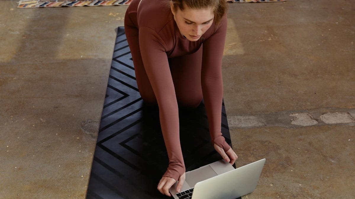 Woman in yoga attire kneeling on mat using laptop indoors, blending fitness with technology.