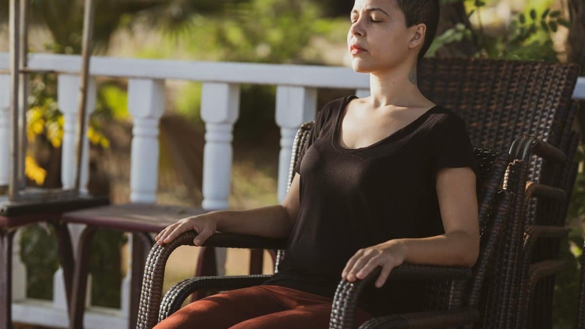 A woman in meditation with eyes closed sitting on a chair outdoors in the sunlight.