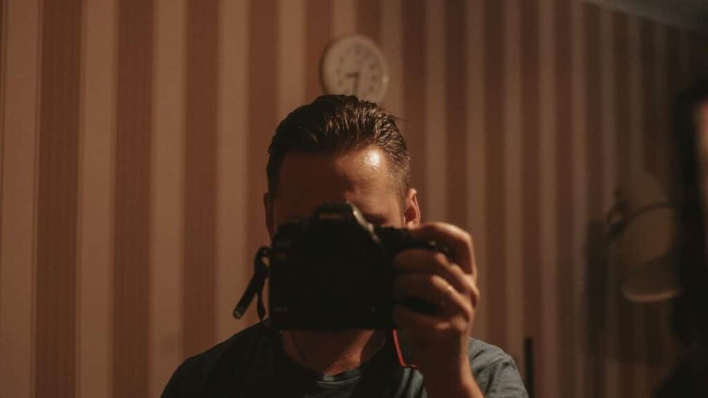 A man holding a camera, capturing his reflection in a dimly lit room with striped wallpaper.