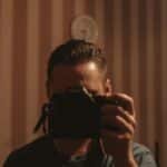 A man holding a camera, capturing his reflection in a dimly lit room with striped wallpaper.
