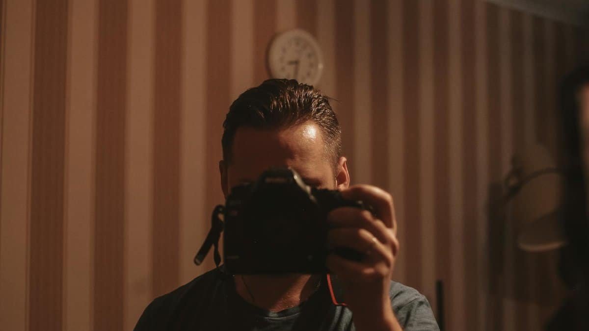 A man holding a camera, capturing his reflection in a dimly lit room with striped wallpaper.