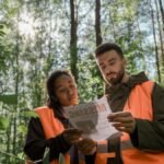 Two forestry workers in safety gear examine a missing person flyer in a sunlit forest.