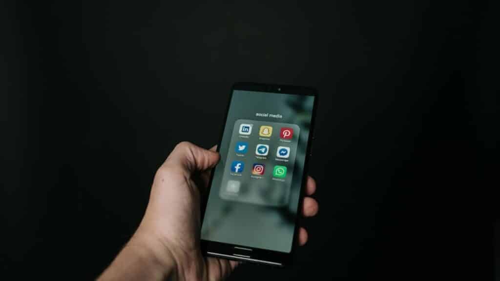 Close-up of a hand holding a smartphone displaying various social media app icons on a dark background.