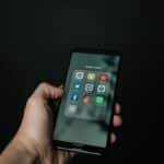 Close-up of a hand holding a smartphone displaying various social media app icons on a dark background.