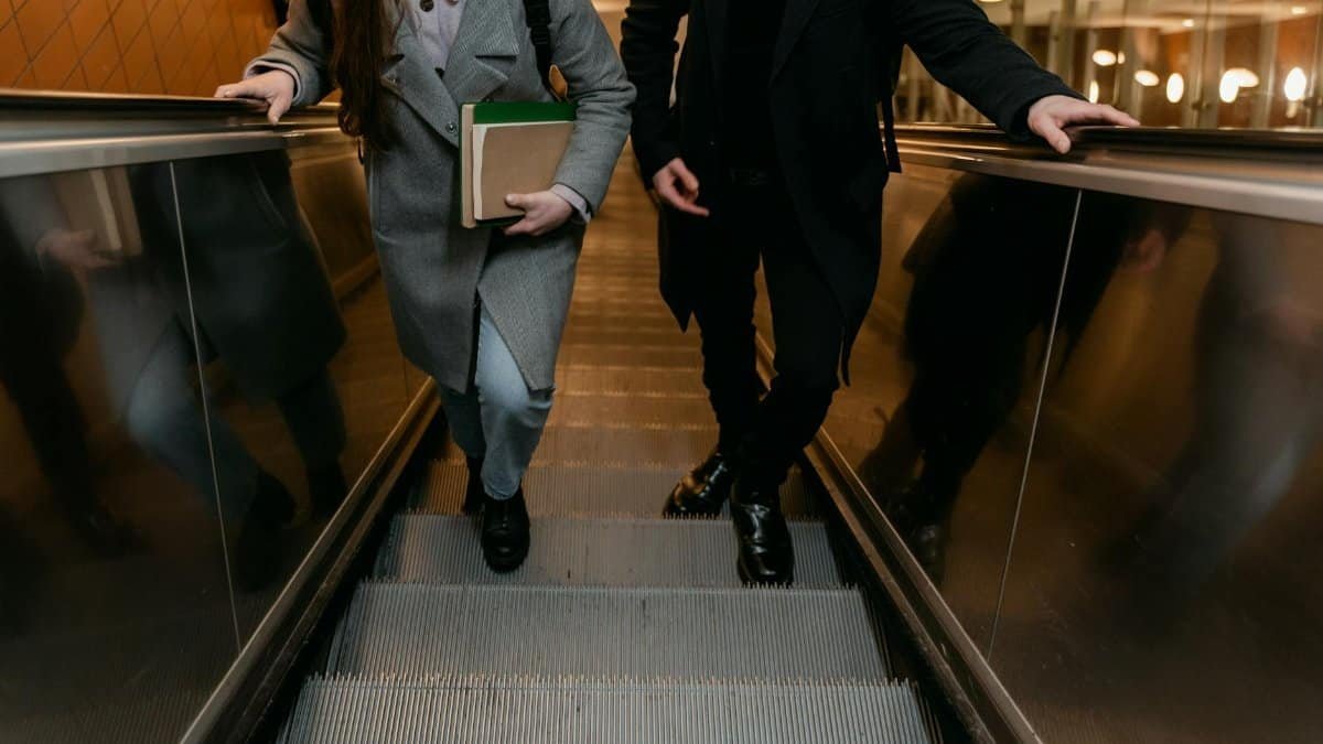 Two adults on an escalator indoors, holding books and bags.