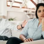 Young bearded Sikh man in casual wear and turban arguing with sad wife sitting on sofa