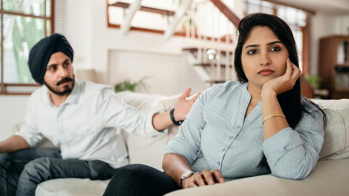 Young bearded Sikh man in casual wear and turban arguing with sad wife sitting on sofa
