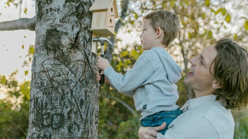 A joyful father assists his son in hanging a birdhouse on a tree, fostering a love for nature.